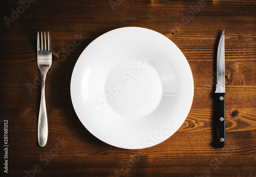 White empty plate with a fork and knife on a wooden table.