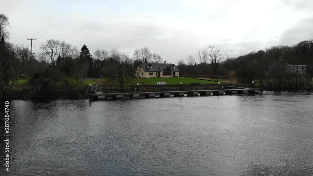 Aerial - Fly through grass field and a river towards a single English house nearby the lake. Aerial - Fly through grass field and a river towards a single English house nearby the lake.