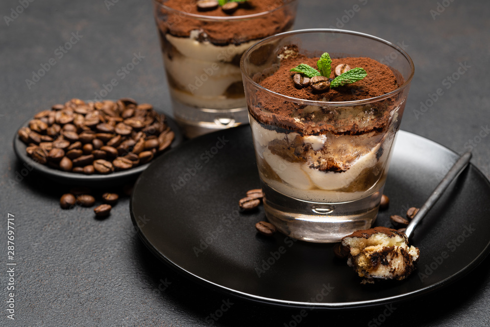Portion of Classic tiramisu dessert in a glass cup on dark concrete background Portion of Classic tiramisu dessert in a glass cup on dark concrete background