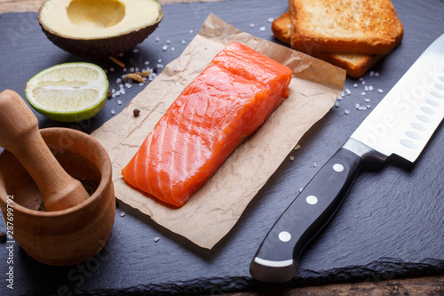 Salmon, avocado, lime and toasts laying with knife on slate background.