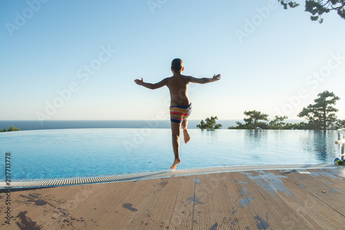 Boy jumping in swimming pool at sunset. European boy in colorful swimming trunks is running to jump into swimming pool during sunset with sea line on the background. Jumping set