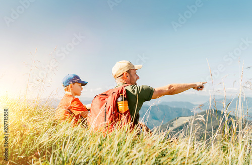 Father showing something interesting to his teenager son sitting on the grass during their mounting hiking walking.