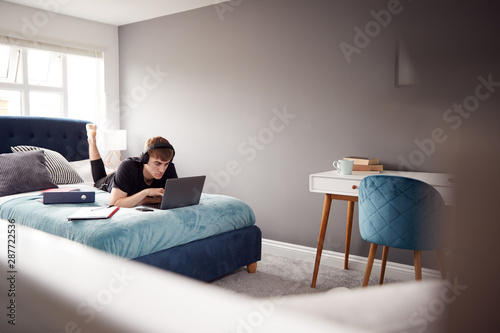Male College Student Wearing Headphones Lies On Bed In Shared House Working On Laptop © Monkey Business
