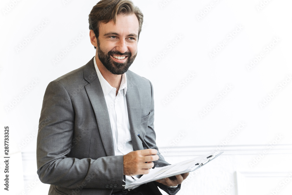 Image of bearded joyful businessman looking forward and holding clipboard while working in office Image of bearded joyful businessman looking forward and holding clipboard while working in office
