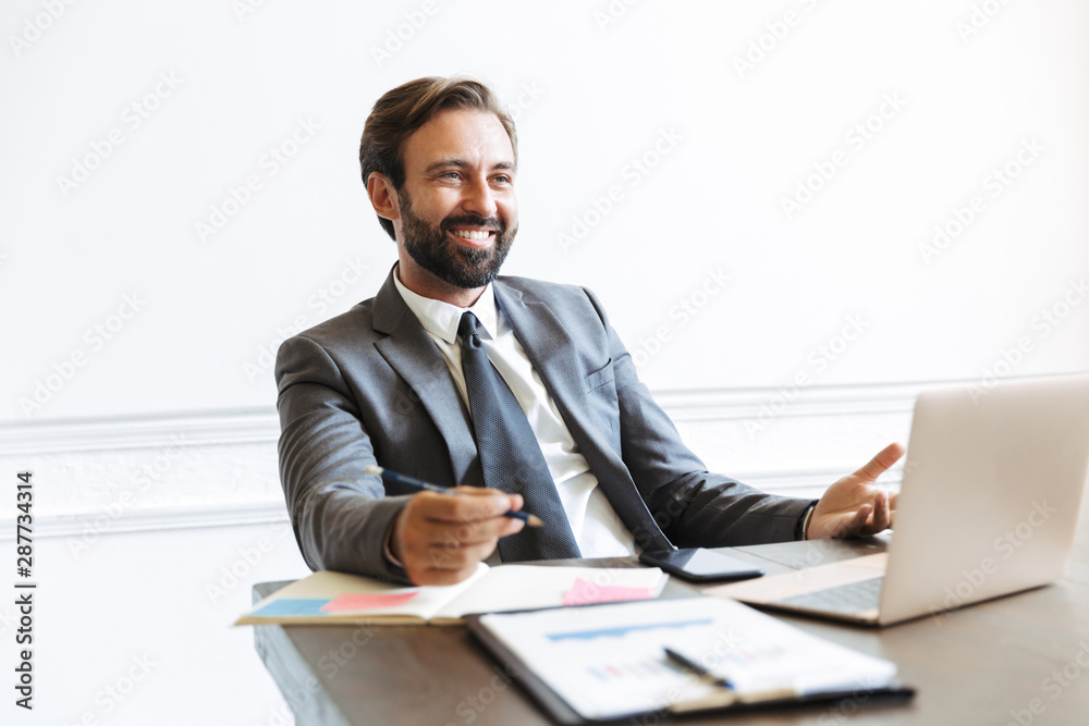 Image of joyful handsome businessman working at laptop in office while looking forward at copyspace Image of joyful handsome businessman working at laptop in office while looking forward at copyspace