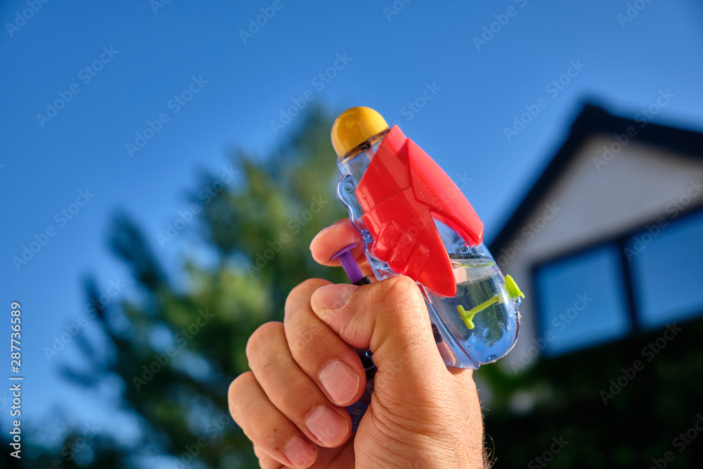 A colorful water pistol in a caucasian male hand in front of  a house and the blue sky of a sunny summer day