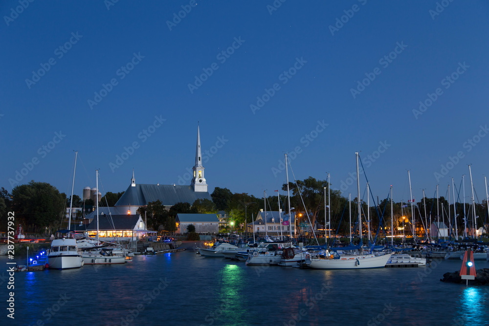 Saint-Michel-de-Bellechasse marina seen during an early evening summer blue hour, with sailboats, historical 1873 church and waterside restaurant in the background, Quebec, Canada