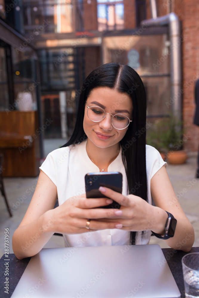 Charming business woman online ordering via mobile phone while sitting in coffee shop during recreation time. Female student chatting on cell telephone, sitting near college