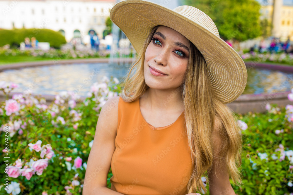 Young charming Caucasian blonde woman in fashionable wicker hat dreaming about something good while sitting alone in park in summer day during leisure time. Thoughtful female enjoying recreation