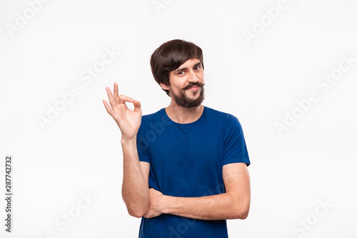 Handsome brunet bearded man with mustaches in a blue shirt showing gesture of OK sign standing isolated over white background. OK gesture.