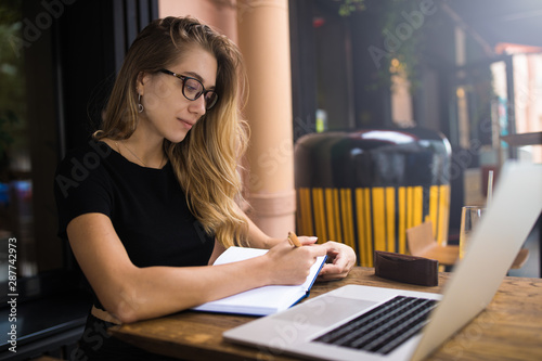 Female successful entrepreneur writing information in textbook while sitting with laptop computer outside restaurant in summer day during recreation time. Woman clever student having online education