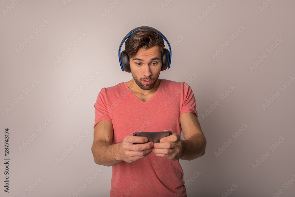 Hipster guy with wireless headphones on head playing games on cell telephone while standing isolated in studio against wall with copy space Hipster guy with wireless headphones on head playing games on cell telephone while standing isolated in studio against wall with copy space