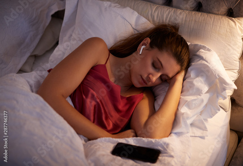 Woman Sleeping In Bed Wearing Wireless Earphones Connected To Mobile Phone photo