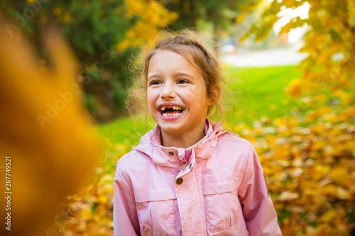 Portrait of Cute little girl with missing teeth playing with yellow fallen leaves in autumn forest. Happy child laughing and smiling. Sunny autumn forest  sun beam. 