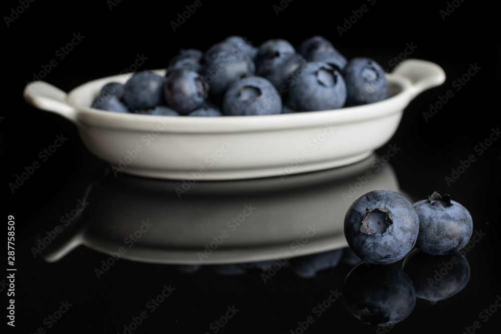 Lot of whole fresh blue bilberry in white oval ceramic bowl isolated on black glass
