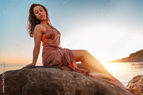 Beautiful tanned woman in underwear sitting on the coastal rocks. Sea and sunset in the background. Copy space © _KUBE_