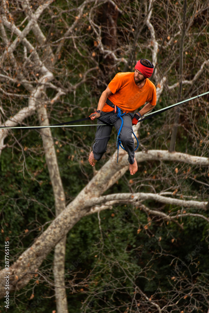 Naklejka premium A man is walking along a stretched sling. Highline in the mountains. Man catches balance. Performance of a tightrope walker in nature. Highliner on the background of valley.