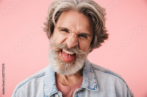 Portrait of amusing old man with gray beard grimacing and sticking out his tongue at camera