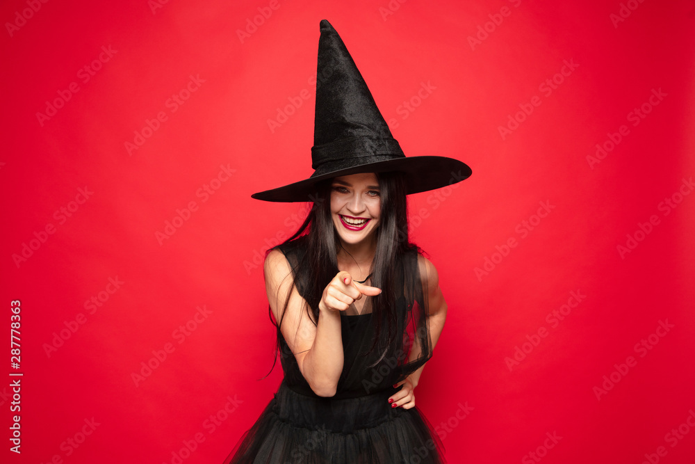 Young brunette woman in black hat and costume on red background. Attractive caucasian female model. Halloween, black friday, cyber monday, sales, autumn concept. Copyspace. Pointing, showing, choosing Young brunette woman in black hat and costume on red background. Attractive caucasian female model. Halloween, black friday, cyber monday, sales, autumn concept. Copyspace. Pointing, showing, choosing