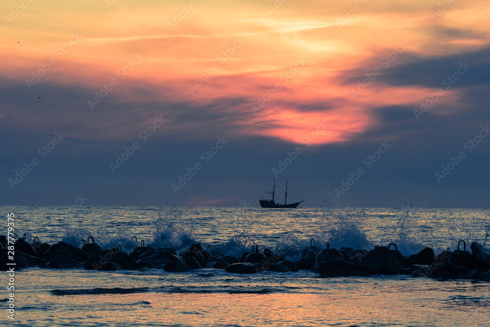 Colorful landscape with a ship sailing on the sea and waves crashing the breakwater under the dramatic sky during sunset.