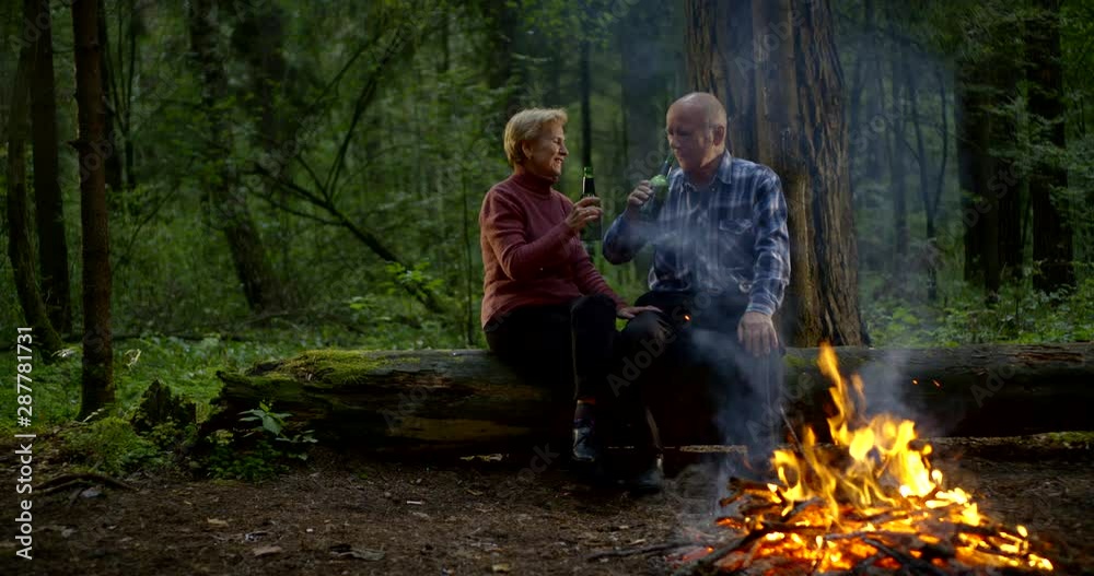 An elderly love couple sit in the evening forest on a log by the fire and drink a bottle of beer, talk, shrug off the smoke. An elderly love couple sit in the evening forest on a log by the fire and drink a bottle of beer, talk, shrug off the smoke.