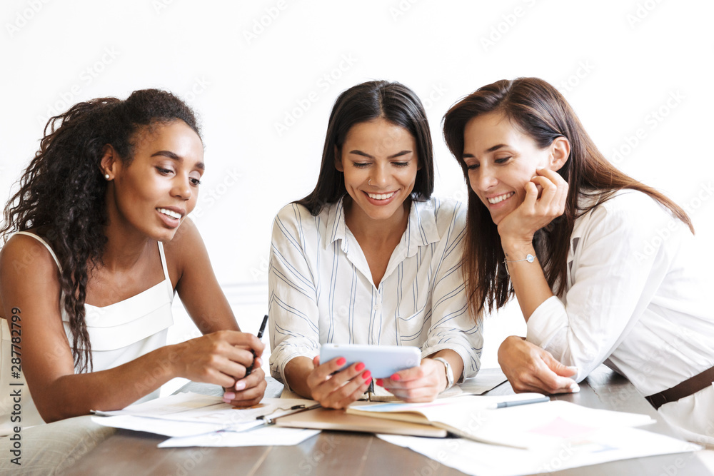 Cheery young business women colleagues in office indoors using mobile phone talking with each other. Cheery young business women colleagues in office indoors using mobile phone talking with each other.