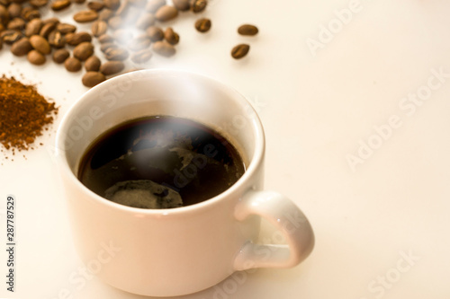 White cup of espresso with coffee grains and ground coffee background. International Coffee Day. White background with copy space for text. Steam from a cup of coffee