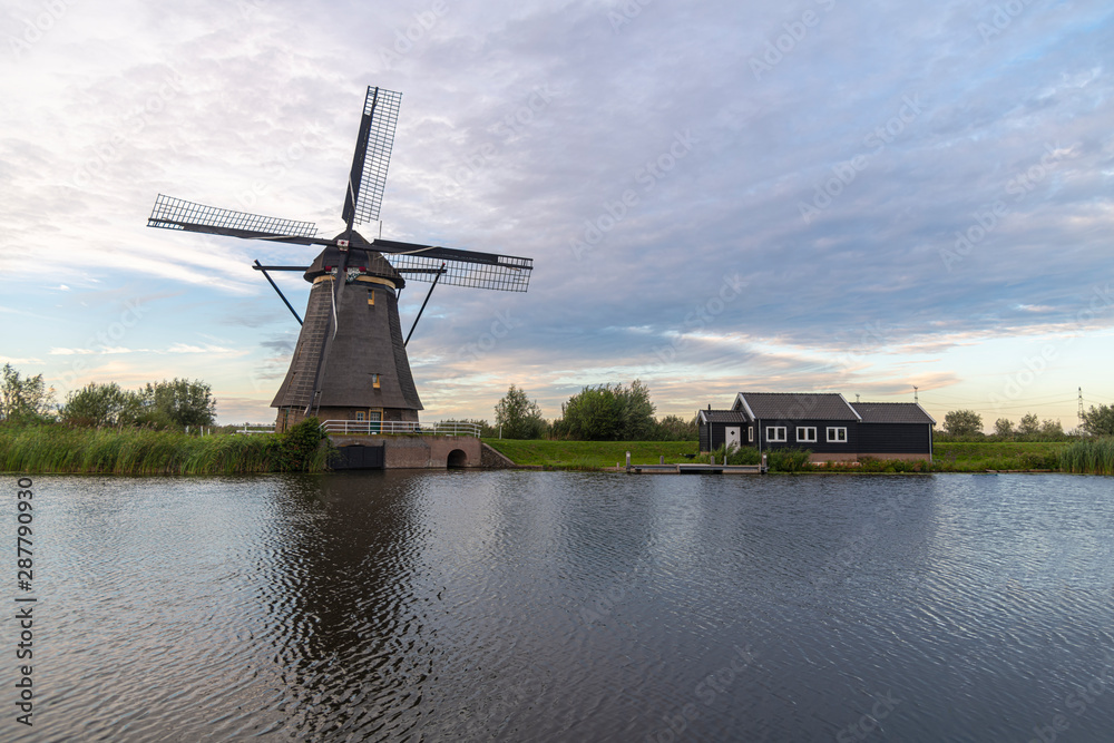 Dutch windmill laying along the canal with wild grass blown by strong winds at the early sunset moment