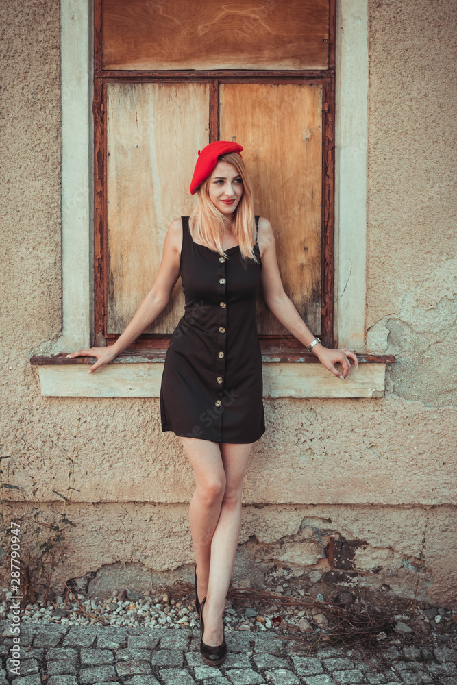 Attractive blond woman in a stylish summer outfit with high heels, black  dress and red French beret posing in front of large historic wooden windows  in town in summer sunshine Stock Photo