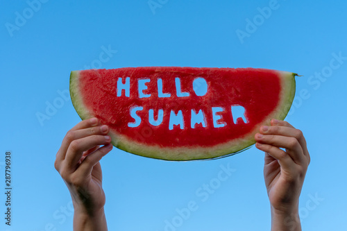 A piece of watermelon against a blue sky. Children's hands are holding a slice of watermelon with the text Hello Summer. Summer time concept