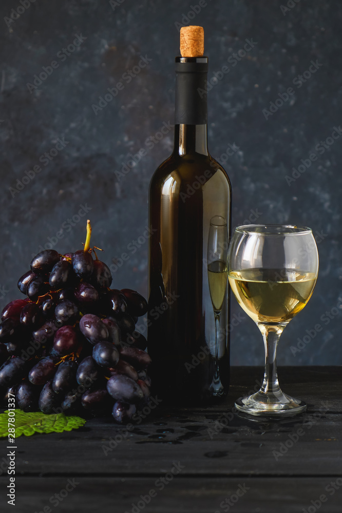 Black grapes, a glass of white wine and a bottle of wine on dark table