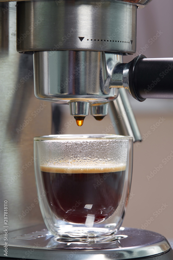 Freshly made espresso in a beautiful transparent double-circuit mug is on the coffee maker. Close up, vertical. Household coffee makers.