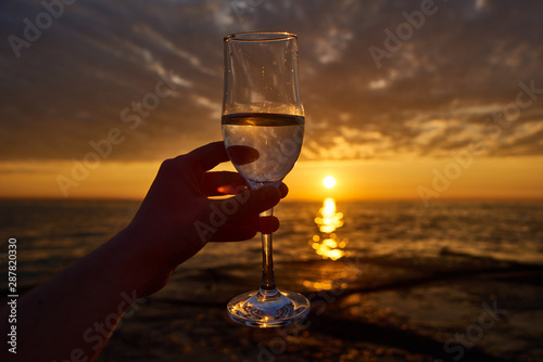 hand holding glass of white wine and with sea and beautiful sunset at background, close-up. Summer evening relaxed mood concept