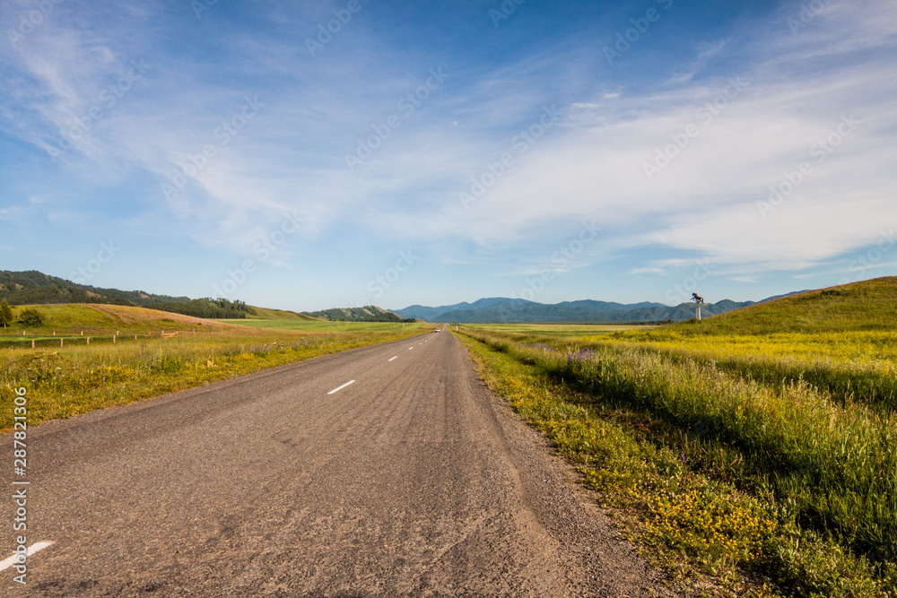 Altai Republic. The road between the hills in the green steppe. Mountain ranges are visible on the horizon.Russia. Altai Republic. The road between the hills in the green steppe. Mountain ranges are visible on the horizon.Russia.