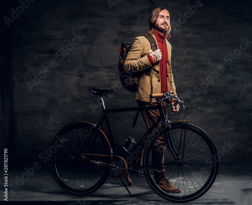 Trendy hipster in jacket is standing at dark photo studio with his retro bike.