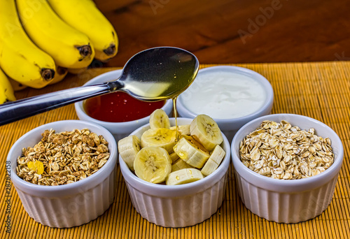 Banana sliced on ramekin and spoon pouring honey with oatmeal, granola, plain yogurt and honey as side dishes under bamboo mat with bunch of bananas in the background © Pedro Turrini Neto