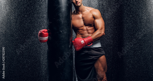 Young muscular man in red boxer gloves is posing with punching bag at dark room with water splashes on the wall. © Fxquadro