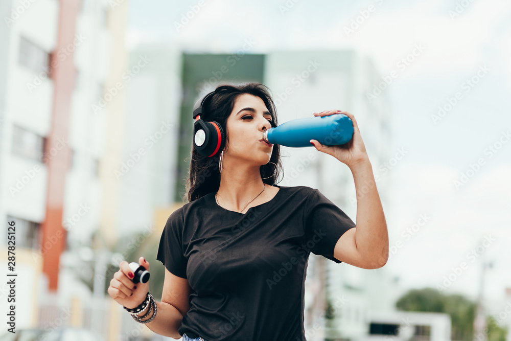 Beautiful young woman using headphone listening to music and drinking water outdoors