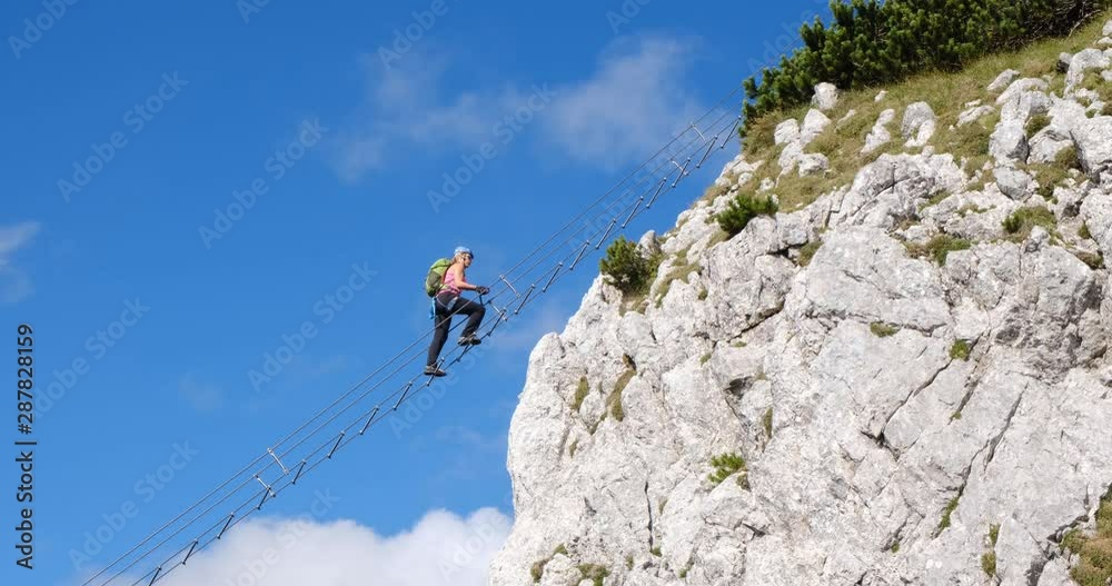 Woman switching lanyards and carabiners on a via ferrata ladder suspended high in the air, against blue sky and white clouds. Location: Donnerkogel mountain near Gosau.