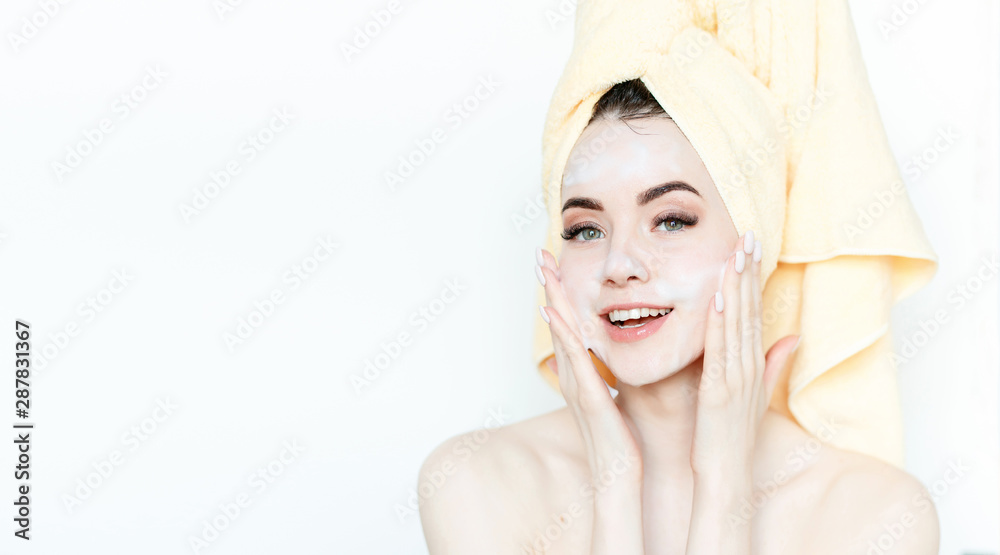 Fototapeta premium Portrait of cheerful woman applying foam for washing on her face. Beautiful model with towel on head with healthy glow perfect smooth skin. Young girl enjoys facial skincare routine.