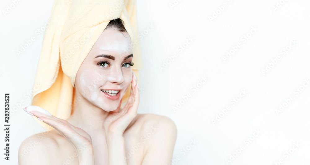Naklejka premium Portrait of cheerful woman applying foam for washing on her face. Beautiful model with towel on head with healthy glow perfect smooth skin. Young girl enjoys facial skincare routine.