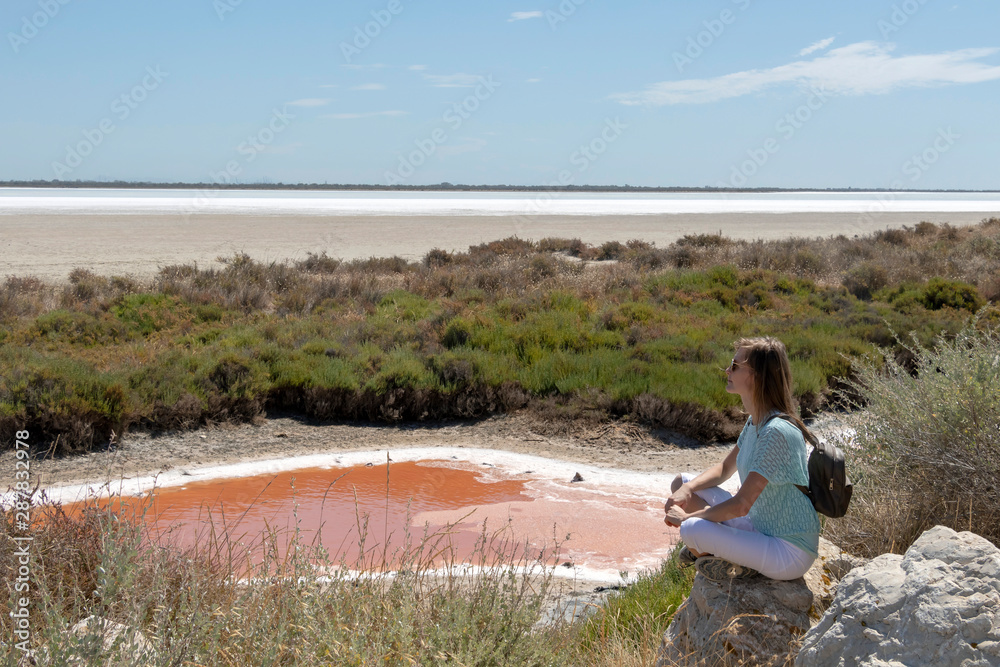 Young blond woman sitting on the rock next to colorful salt lagoon in Camargue Nature Park, Southern France