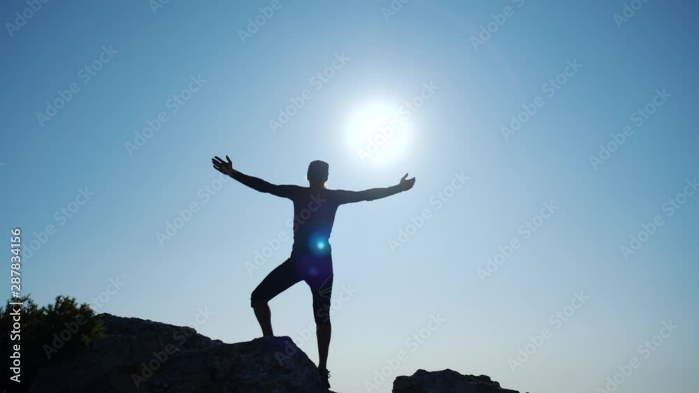 Young long-haired inspired man raises his hands up standing on the top of a mountain above the sea against beautiful blue sky. Silhouette of a happy hiker man standing on the summit. Young long-haired inspired man raises his hands up standing on the top of a mountain above the sea against beautiful blue sky. Silhouette of a happy hiker man standing on the summit.