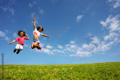 Two happy girls jump high over blue sky on lawn