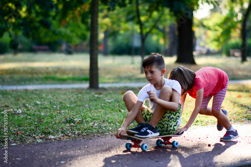 A girl pushes a boy sitting on a skateboard in a park. Brother and sister play in the park while skating in nature. Concept of active life of children in nature.