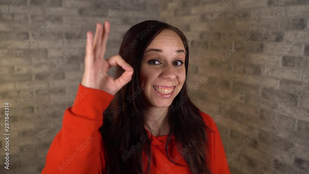 A beautiful young woman in a red hoodie shows is smiling and showing sign of OK on a gray brick background. A beautiful young woman in a red hoodie shows is smiling and showing sign of OK on a gray brick background.