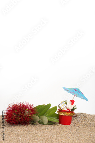 A sprig of red flowering pohutukawa (New Zealand Christmas tree) with Christmas decorations and a cocktail umbrella on sand with a white background.
