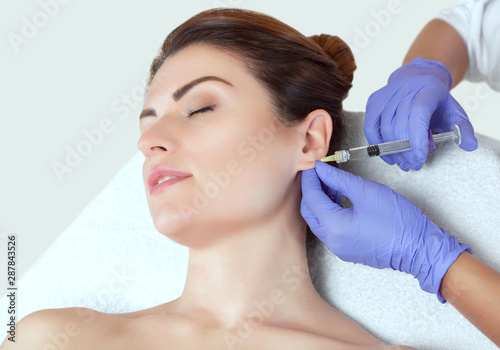 The doctor cosmetologist makes the injections procedure for smoothing wrinkles and against flabbiness of the skin on earlobe of a beautiful, young woman in a beauty salon. photo