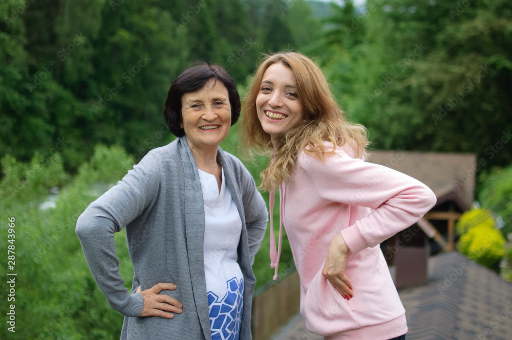 Outdoors portrait of happy smiling mother and daughter who are looking at the camera outside over landscape of forest and mountains.