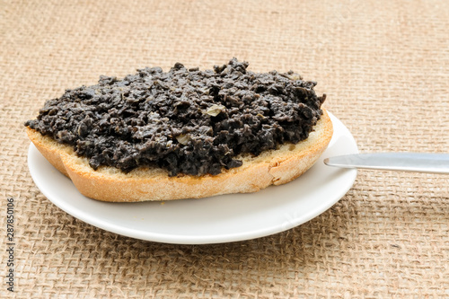 Fresh tapenade sandwich with slice of wheat bread in a white sauser and table knife on a burlap table napkin. Homemade black olive paste with anchovies, garlic and olive oil.